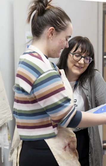 Two people in an art studio discussing a blue printmaking plate, with aprons and art supplies in the background. Two people in an art studio discussing a blue printmaking plate, with aprons and art supplies in the background.